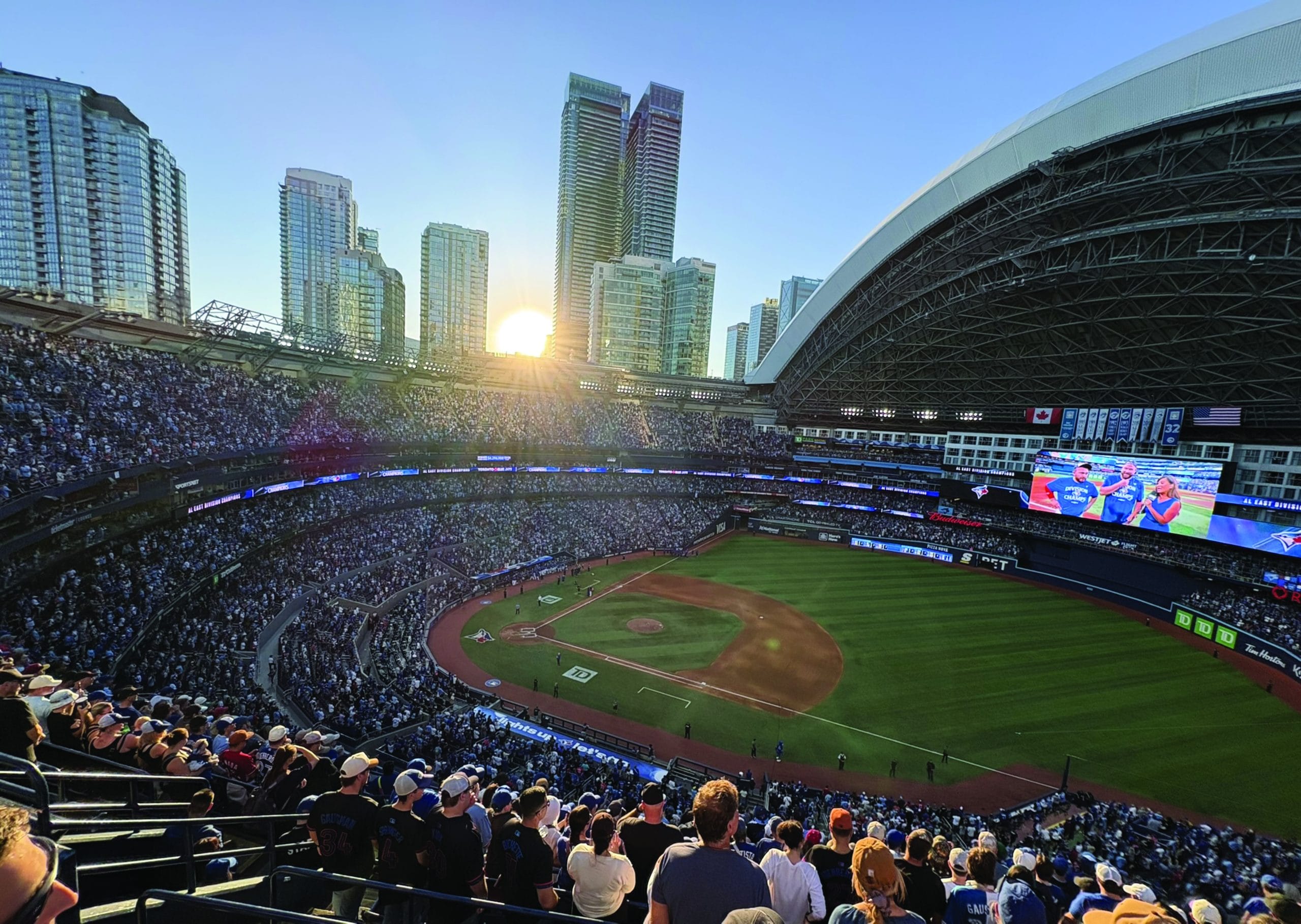 A large crowd watches a baseball game in a stadium with a partially open roof, surrounded by tall city buildings as the sun sets—capturing the excitement of the Wide World Of Sports.