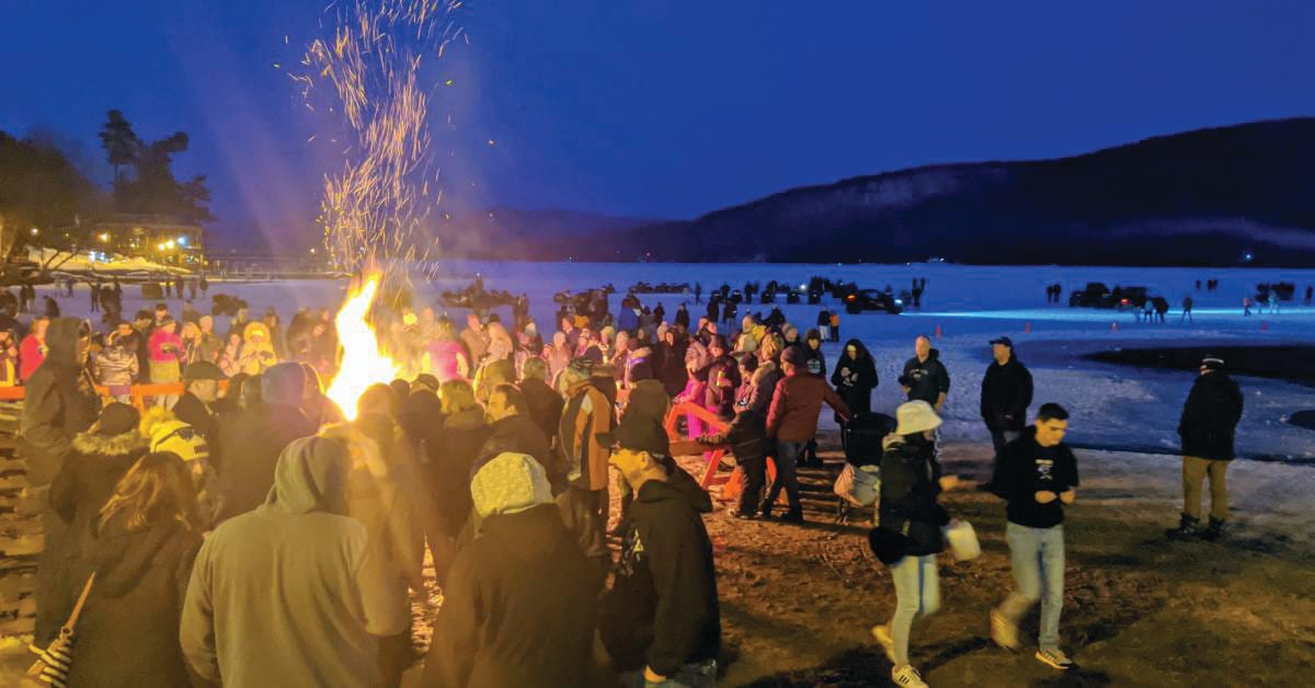 A large group gathers around a bonfire on a beach at dusk, sparks rising into the night sky, while others socialize near a frozen lake and snow-covered mountains—capturing the spirit of True Great Adventures out of doors.
