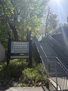 A sunlit entrance to the Walkway Over the Hudson State Historic Park, featuring a large sign and a metal staircase leading upward, surrounded by green trees—a perfect starting point for a Walking Tour of Poughkeepsie.