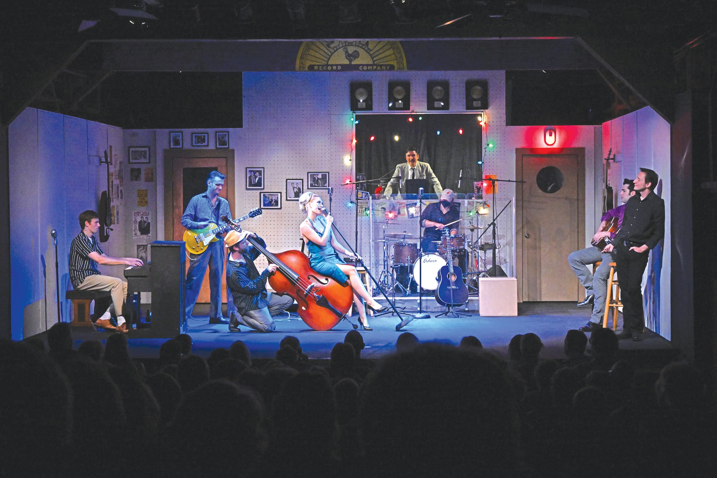 A band performs on stage at Depot Theatre with a pianist, guitarist, upright bass player, singer, and drummer. Two people sit on the right, and lights and framed photos decorate the set. The audience is visible in the foreground.