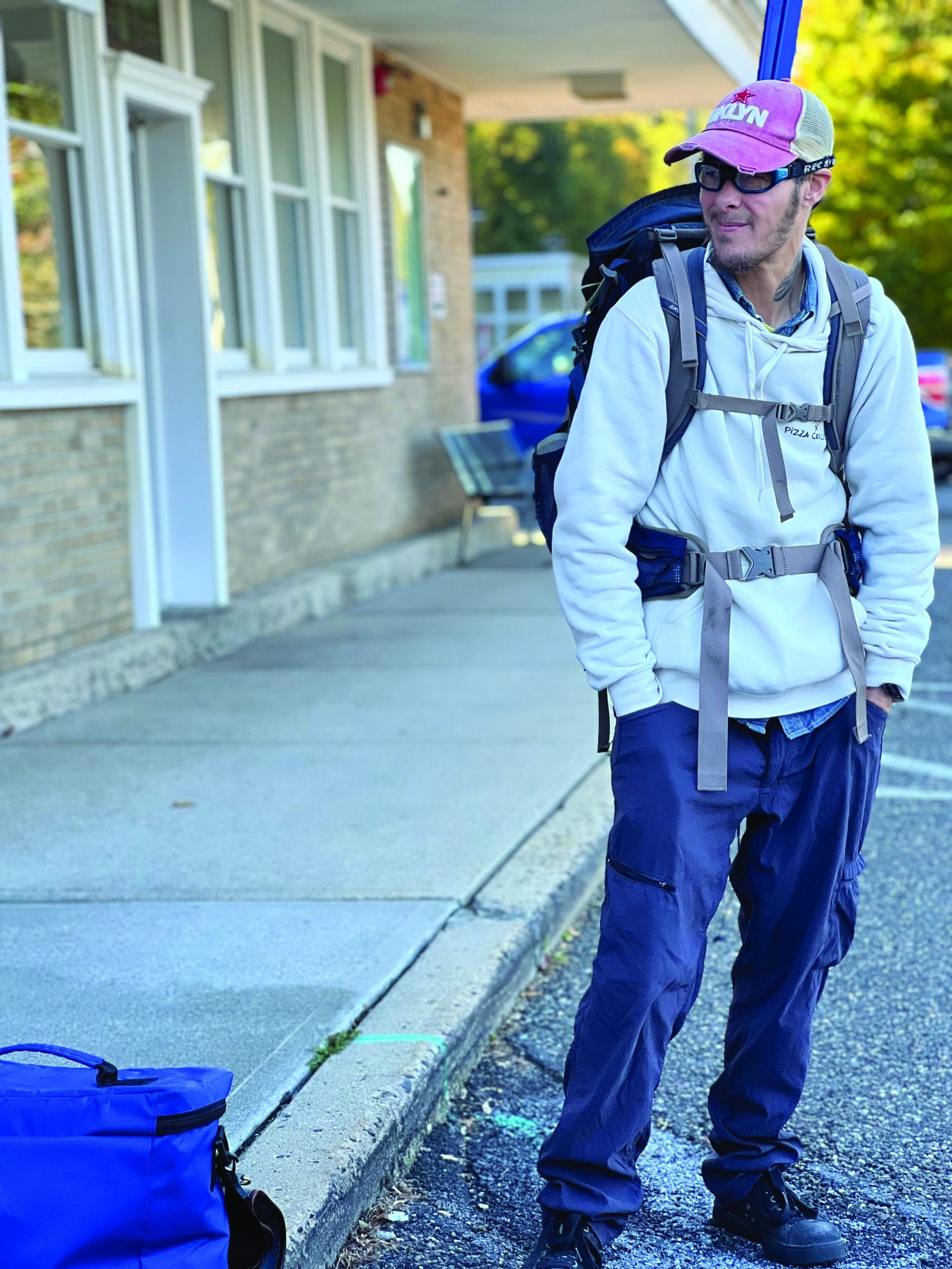 A man wearing a white hoodie, blue cargo pants, a backpack, and a red cap stands on a sidewalk near the Pizza By Rail building, smiling. A blue duffle bag is on the ground near him.