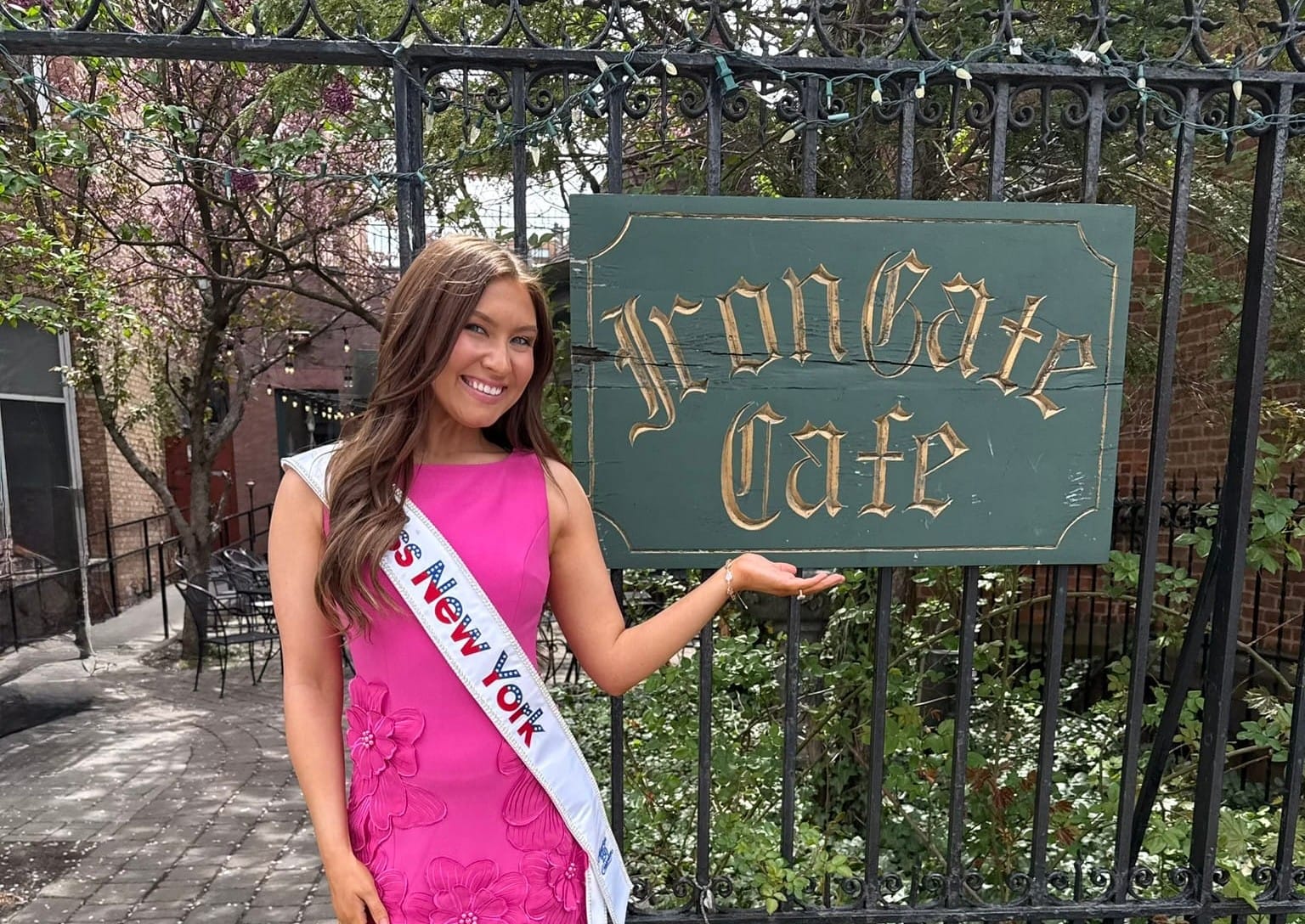 A woman in a pink dress and Miss New York sash smiles and gestures toward a green Iron Gate Cafe sign on a wrought iron fence, as Miss New York Explores Albany NY by Amtrak, with trees and a brick building in the background.