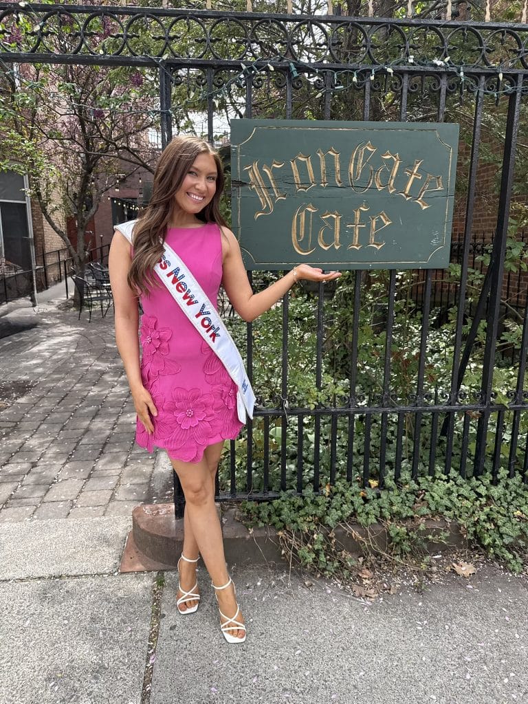 A woman wearing a bright pink dress and a “New York” sash smiles and poses with one hand raised in front of an Iron Gate Cafe sign, standing on a stone path near a black iron fence and greenery.
