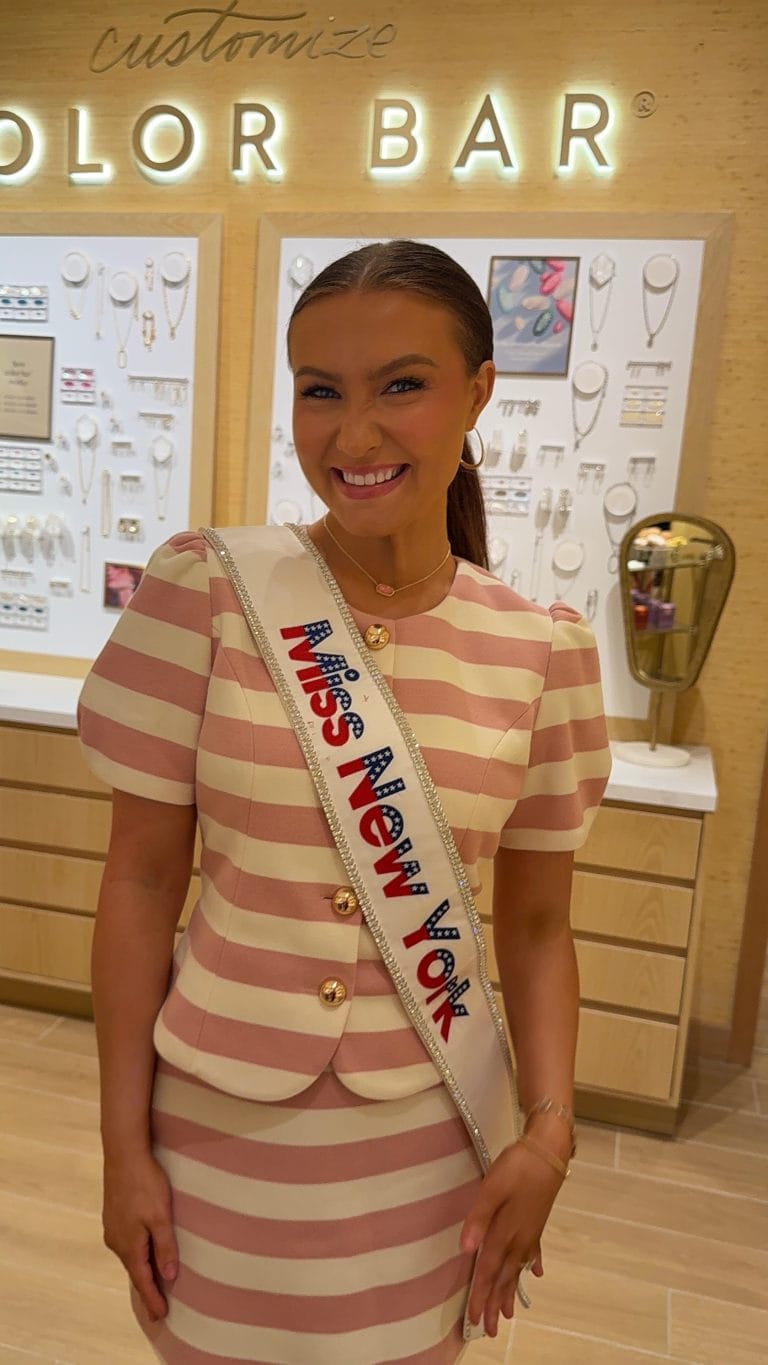 A smiling woman wearing a pink and beige striped dress and a Miss New York sash stands in a jewelry store with display cases and a wall of necklaces behind her.