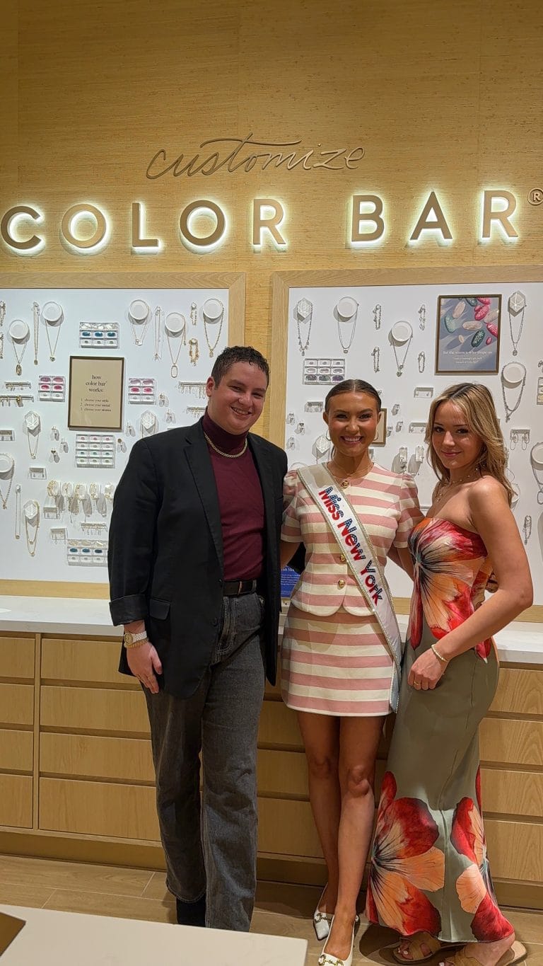 Three people pose and smile in front of a wall display of jewelry under a sign that reads “COLOR BAR.” The woman in the center wears a “Miss New York” sash and a striped dress. The other two wear stylish outfits.