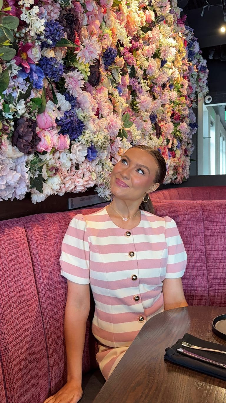 A woman in a pink and white striped dress sits in a red booth, smiling and looking up at a vibrant wall covered in colorful flowers. A set table with black napkins is in front of her.