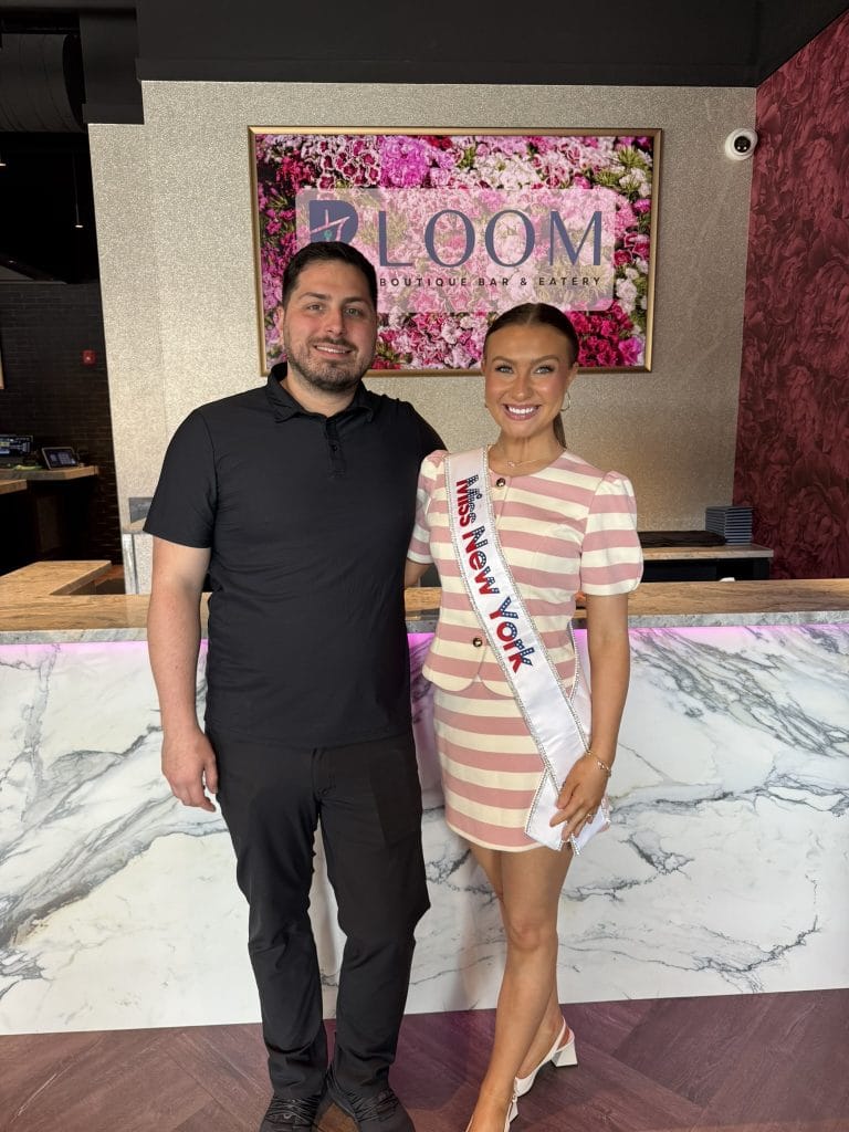 A man in a black shirt stands next to a woman wearing a pink and white striped dress and a Miss New York sash, both smiling in front of a marble counter and a flower sign that says Bloom.