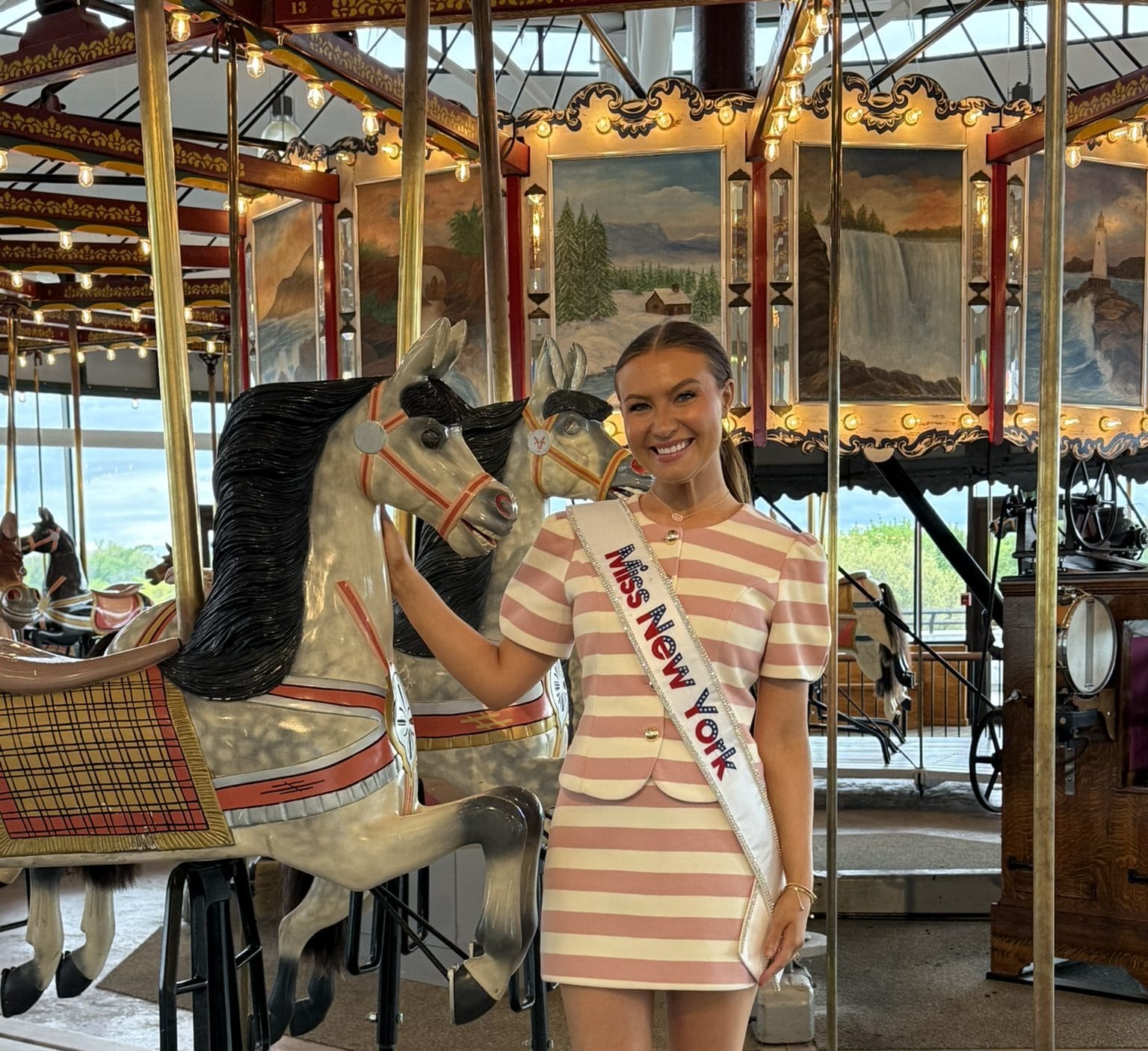 A smiling woman wearing a Miss New York Lake George sash and a pink-and-white striped dress stands next to a carousel horse on a merry-go-round, surrounded by vibrant decorations and painted scenery in the background.