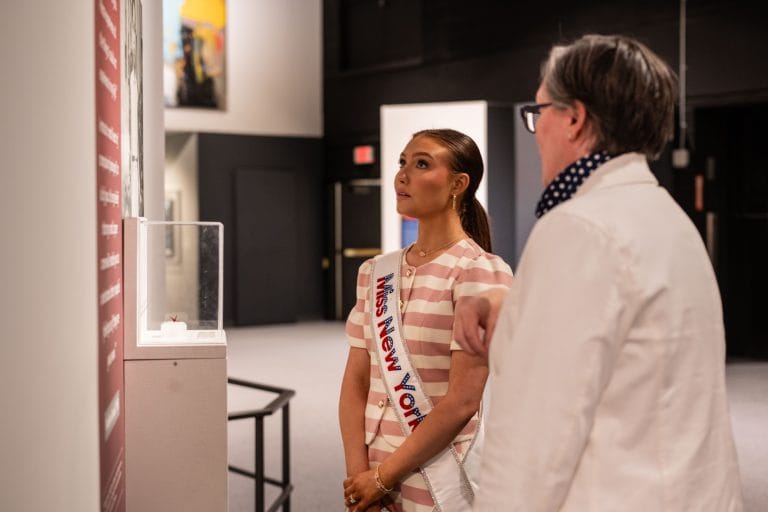 A woman wearing a Miss New York sash stands beside a person in a white jacket, both looking at an exhibit display in a museum or gallery setting.