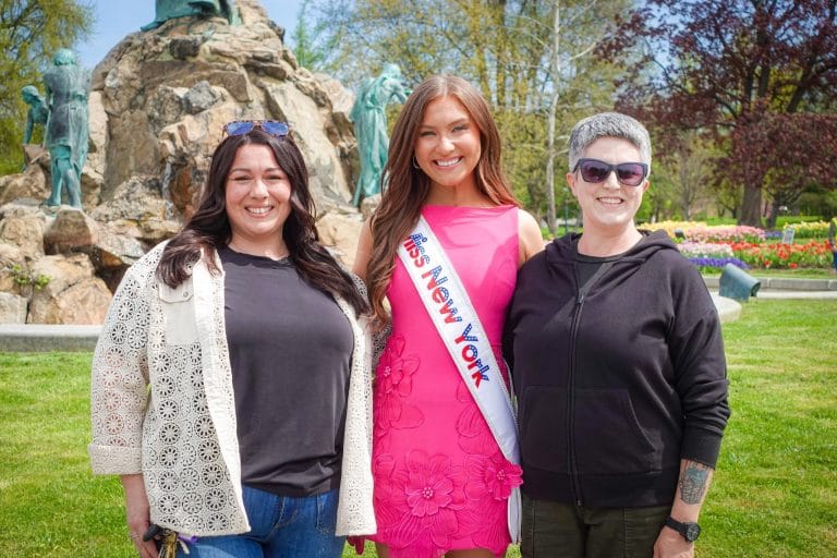 Three women stand together smiling outdoors in a park. The woman in the middle wears a pink dress and a Miss New York sash. The other two women are casually dressed. There are trees and a statue in the background.