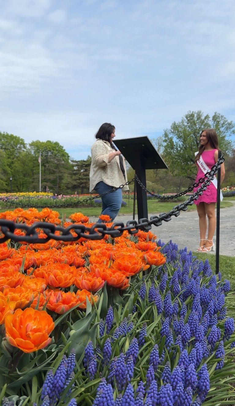 Two women stand by a podium in a garden. One, wearing jeans and a beige top, speaks at the podium; the other, in a pink dress and sash, stands nearby. Bright orange and purple flowers and a black chain line the foreground.