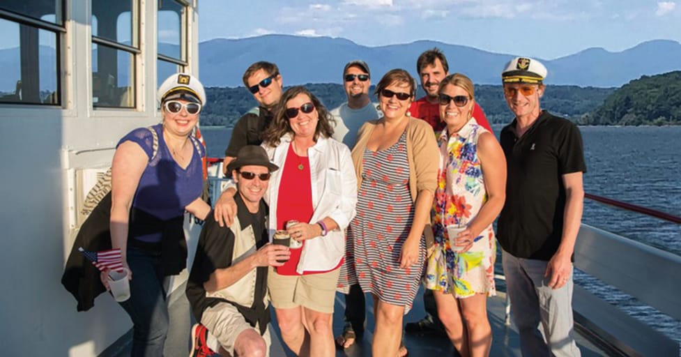 A group of nine adults smiling and posing together on the deck of a boat, with water and mountains in the background on a sunny day. Some are wearing hats and sunglasses, and it looks festive and relaxed.