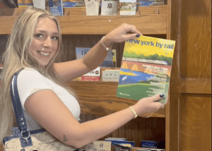 A smiling woman with long blonde hair holds up a New York by Rail magazine in front of a wooden bookshelf filled with various travel brochures, including guides for a Poughkeepsie walking tour.