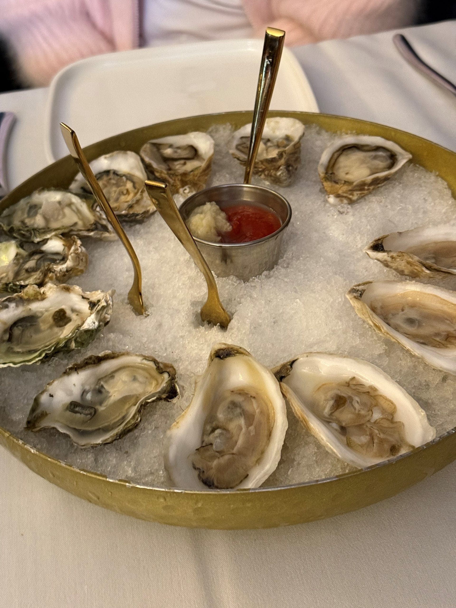 A round tray of raw oysters from Miss New York Lake George rests on crushed ice, served with cocktail sauce and horseradish in the center. Three small forks are among the oysters, with a white plate and napkin in the background.