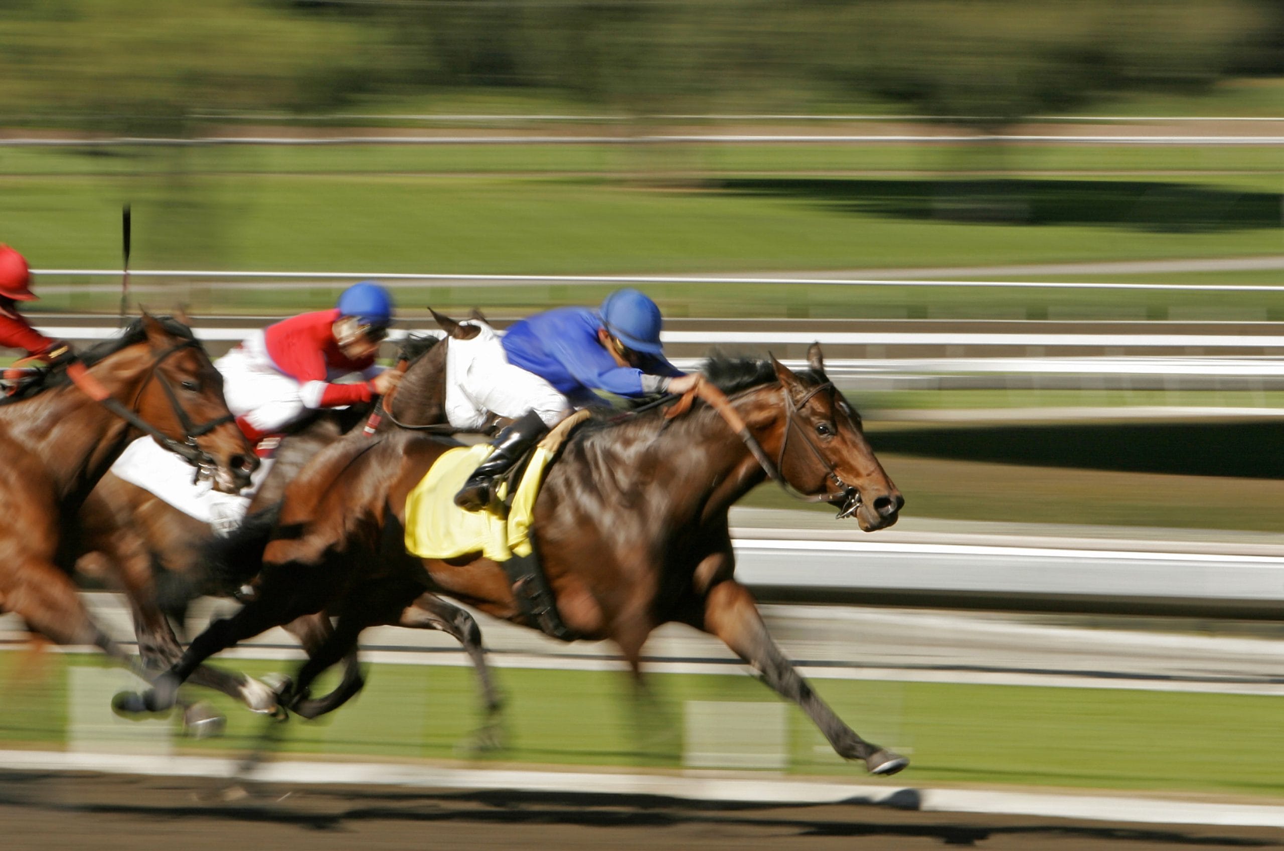 Three jockeys race at full speed on a racetrack during the 2026 Belmont Stakes. The focus is on the leading jockey in blue, while the background and other riders blur to emphasize motion and intensity.