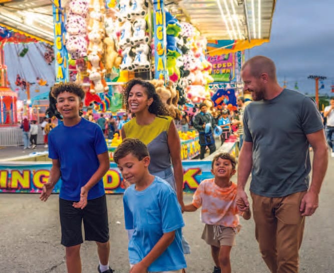 A family of five, including two adults and three children, smiles and walks together at a colorful Syracuse carnival with bright lights and stuffed animal prizes in the background during their city exploring adventure.