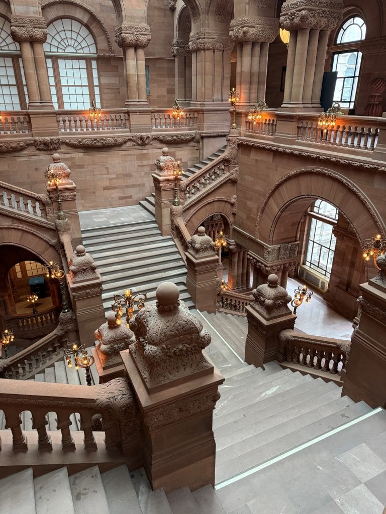 Grand staircase in a historic building with ornate stone railings, arched windows, and decorative columns. Warm lights illuminate the interior, as if preparing for a Miss New York Lake George gala amid these intricate architectural details.