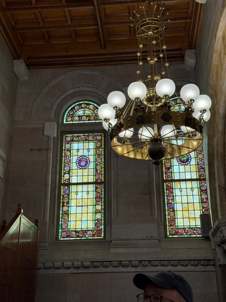 A large ornate chandelier with round lights hangs from a wooden ceiling before two tall stained glass windows in a historic building near Miss New York Lake George. Part of a person in a hat is visible at the bottom right.