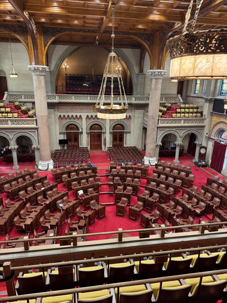 A large, ornate legislative chamber with red carpet, curved rows of brown chairs, high arches, columns, and chandeliers—like a grand hall fit for Miss New York Lake George—viewed from an upper balcony with empty yellow seats.