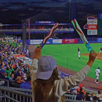 A young girl in a white cap cheers at a Syracuse baseball game, waving two colorful batons in the air while standing near the railing as fans watch from the stands, exploring the excitement of an unforgettable night.