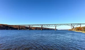 A long steel bridge with multiple supports spans across a wide river under a clear blue sky, with wooded hills on both sides—an iconic sight on a walking tour of Poughkeepsie.