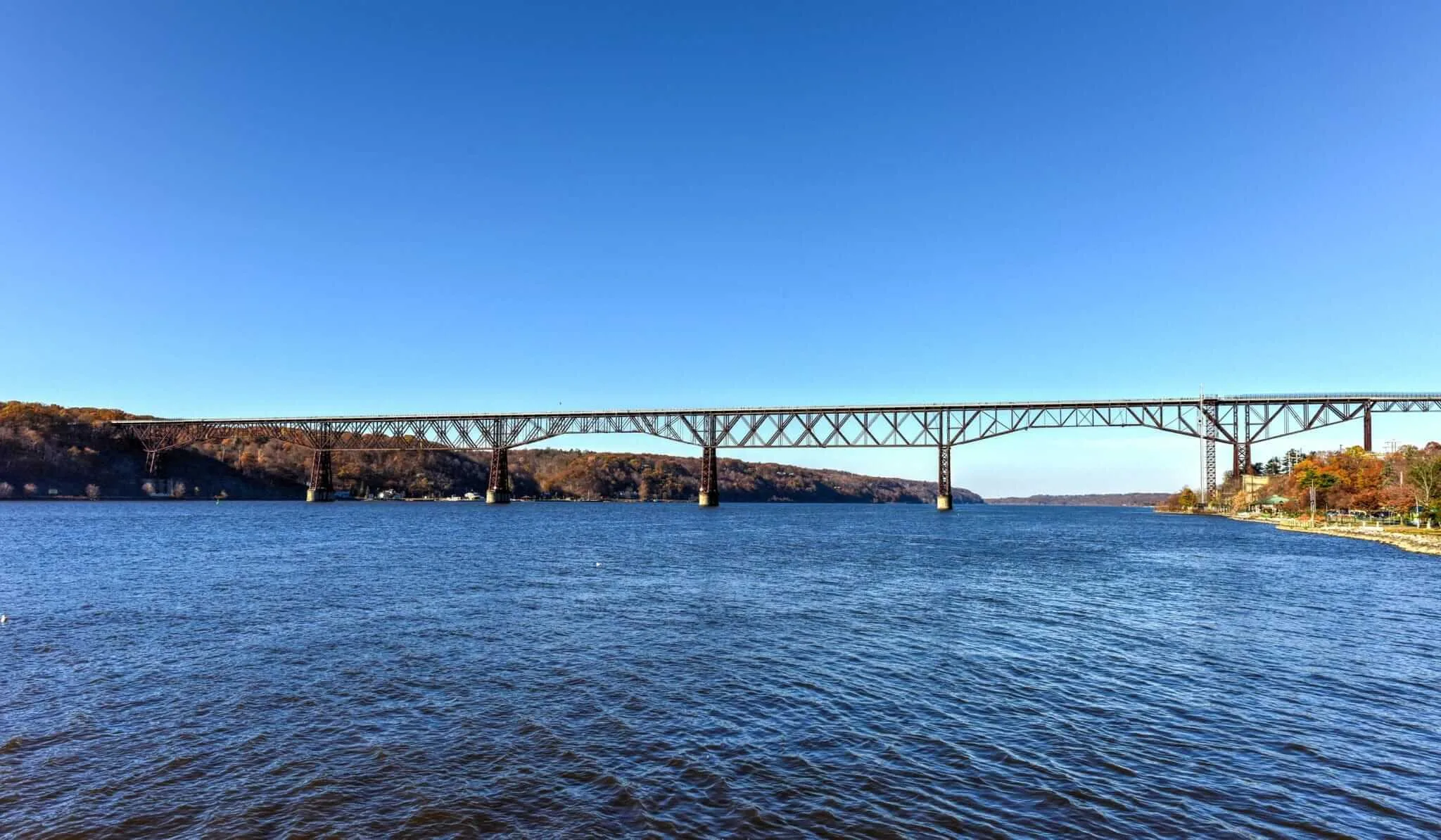 A long steel bridge with multiple supports spans across a wide river under a clear blue sky, with wooded hills on both sides—an iconic sight on a walking tour of Poughkeepsie.