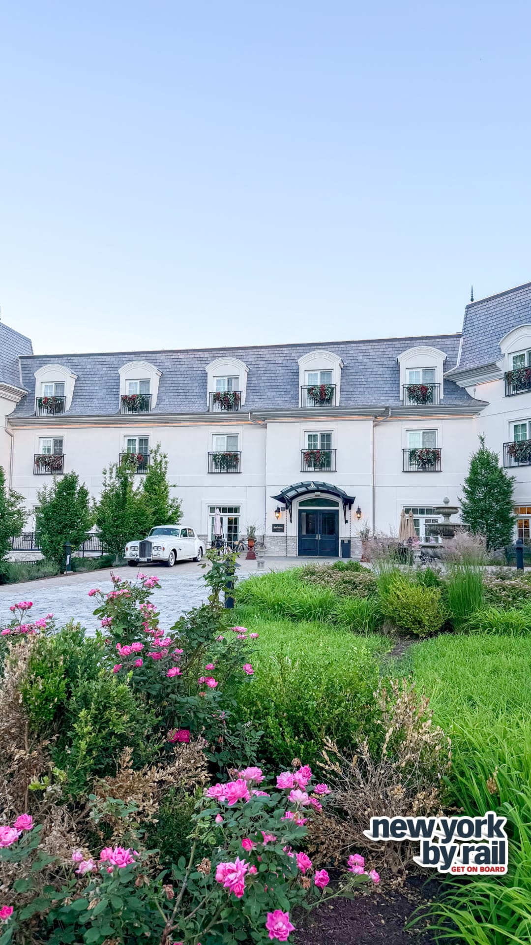 A grand, white, three-story hotel—Miss NY Mirbeau Inn—with dormer windows and flower boxes stands amid lush gardens dotted with pink flowers. A car is parked near the entrance. The clear, light blue sky frames the New York by Rail logo.