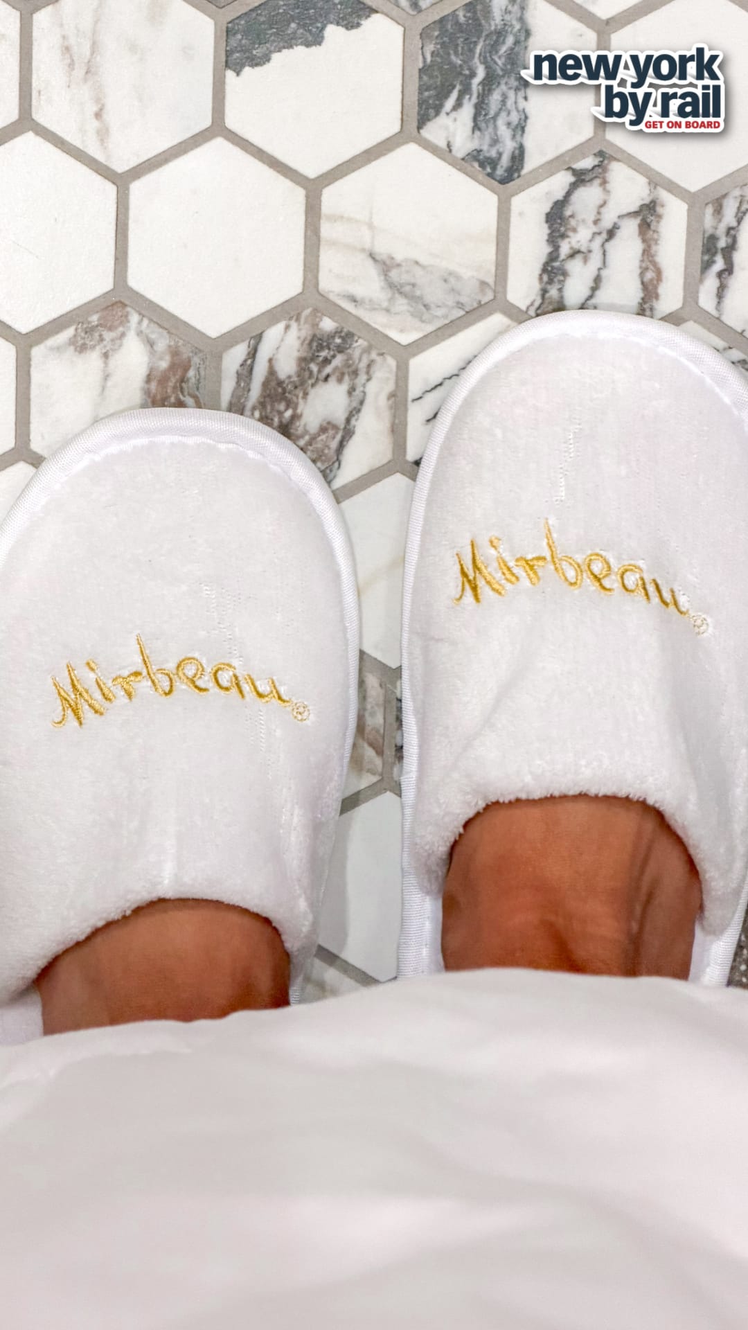 Two feet in white slippers with Mirbeau embroidered in gold, standing on a hexagonal tiled floor at Miss NY Mirbeau Inn. A New York by Rail logo appears in the top right corner.