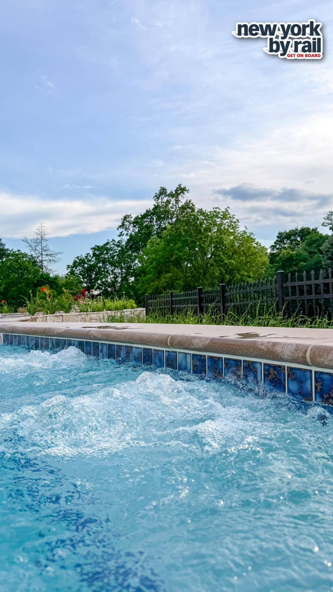 Bubbling water in a blue-tiled hot tub outdoors at Miss NY Mirbeau Inn, surrounded by greenery, flowers, and a black iron fence under a partly cloudy sky. New York By Rail logo appears in the top right corner.