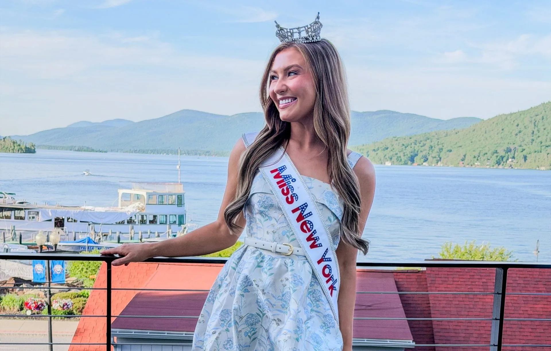 A young woman wearing a tiara, a sash reading Miss New York, a short light blue dress, and white heels stands on a balcony with scenic lake and mountains—Miss New York explores Albany NY by Amtrak.
