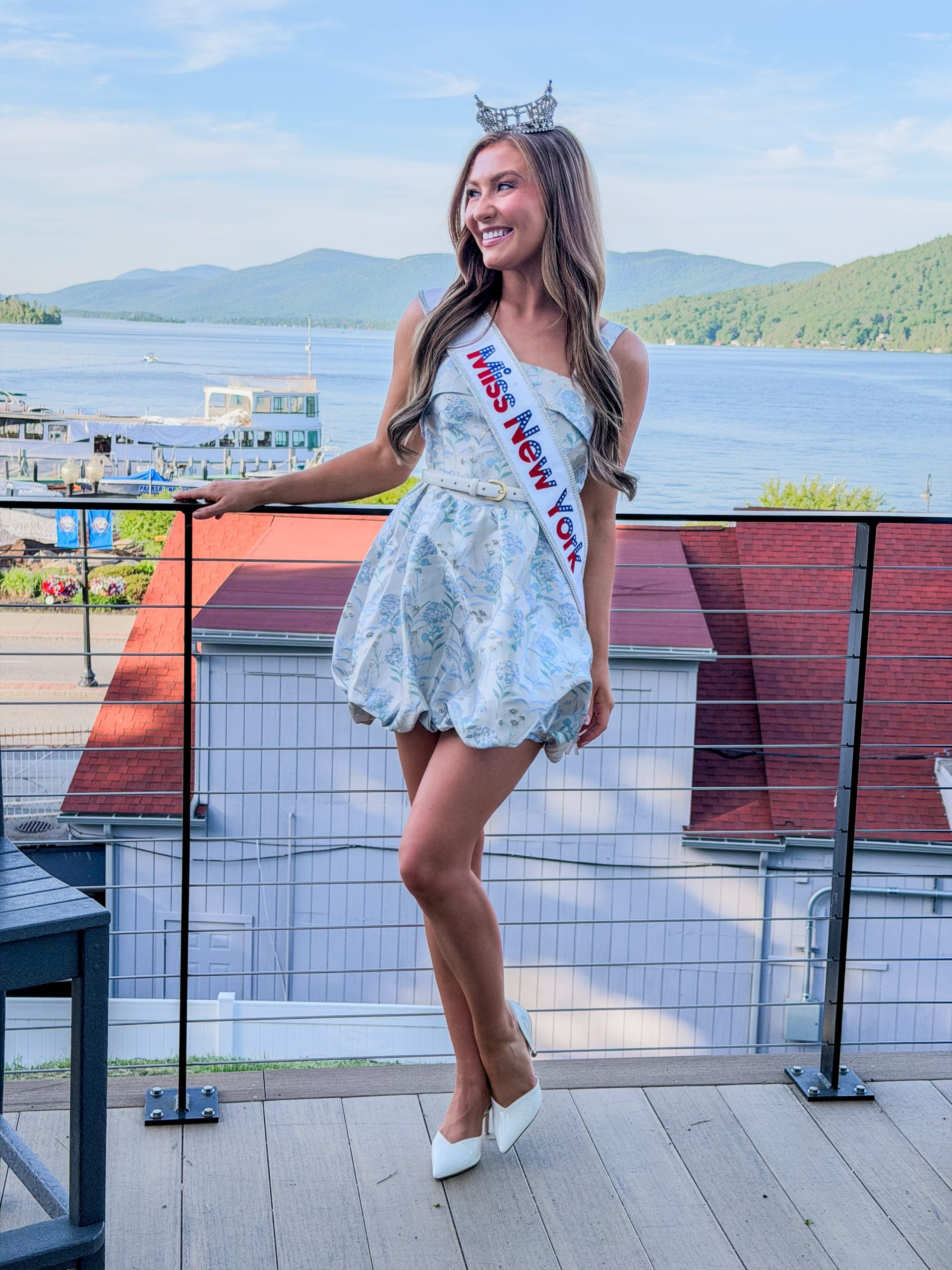 A young woman wearing a tiara, a sash reading Miss New York, a short light blue dress, and white heels stands on a balcony with scenic lake and mountains—Miss New York explores Albany NY by Amtrak.