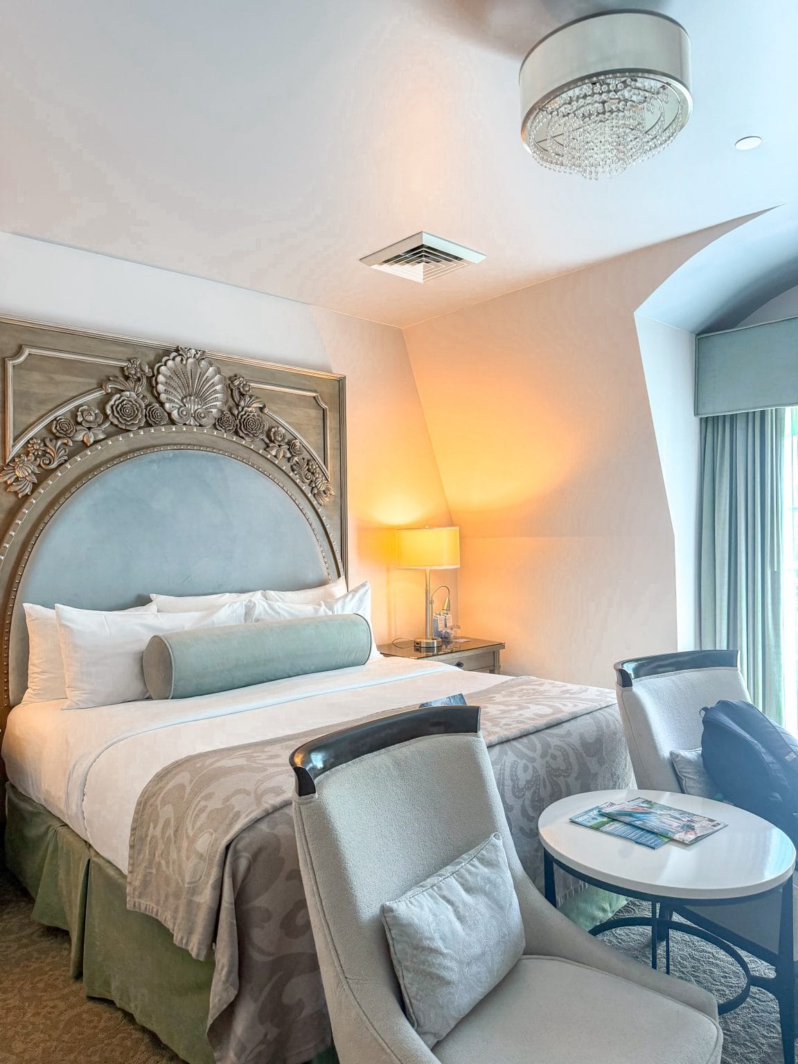 A bright hotel room with a large, ornate headboard, a neatly made bed with white and beige bedding, two armchairs around a small round table, a lamp on a nightstand, and light streaming in from a window.