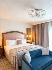 A stylish Miss NY Mirbeau Inn hotel room features a large bed with a plush headboard, patterned bedding, a blue velvet loveseat at the foot, elegant pendant lights, a chandelier, and beige curtains covering large windows.