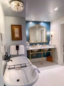 A stylish bathroom at Miss NY Mirbeau Inn features a vintage clawfoot bathtub, white towels, a marble double sink with gold fixtures, a large ornate mirror, blue accent wall, framed artwork, and a chandelier.