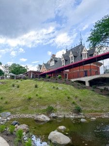 A large, red-brick building with pointed gables and ornate trim sits atop a grassy hill—much like the Miss NY Mirbeau Inn—with a covered walkway and a small stream with rocks in the foreground under a partly cloudy sky.