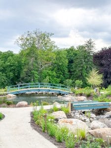 A scenic garden at Miss NY Mirbeau Inn features a winding gravel path, blooming plants, large rocks, a small pond, and two blue wooden footbridges surrounded by lush green trees under a cloudy sky.