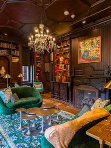 Luxurious library at Miss NY Mirbeau Inn with dark wood paneling, green velvet sofas, a chandelier, bookshelves filled with books, a fireplace, colorful artwork above the mantel, and a patterned green rug on the floor.