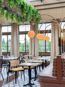 A stylish restaurant interior at Miss NY Mirbeau Inn features hanging plants, globe pendant lights, patterned floor tiles, wicker chairs, and large windows revealing a green outdoor view with striped umbrellas.