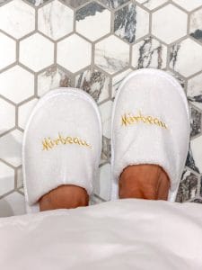 Person wearing a white robe and white slippers with Mirbeau embroidered in gold, standing on a hexagonal tile floor at the Miss NY Mirbeau Inn, surrounded by a mix of white and marble-patterned tiles.