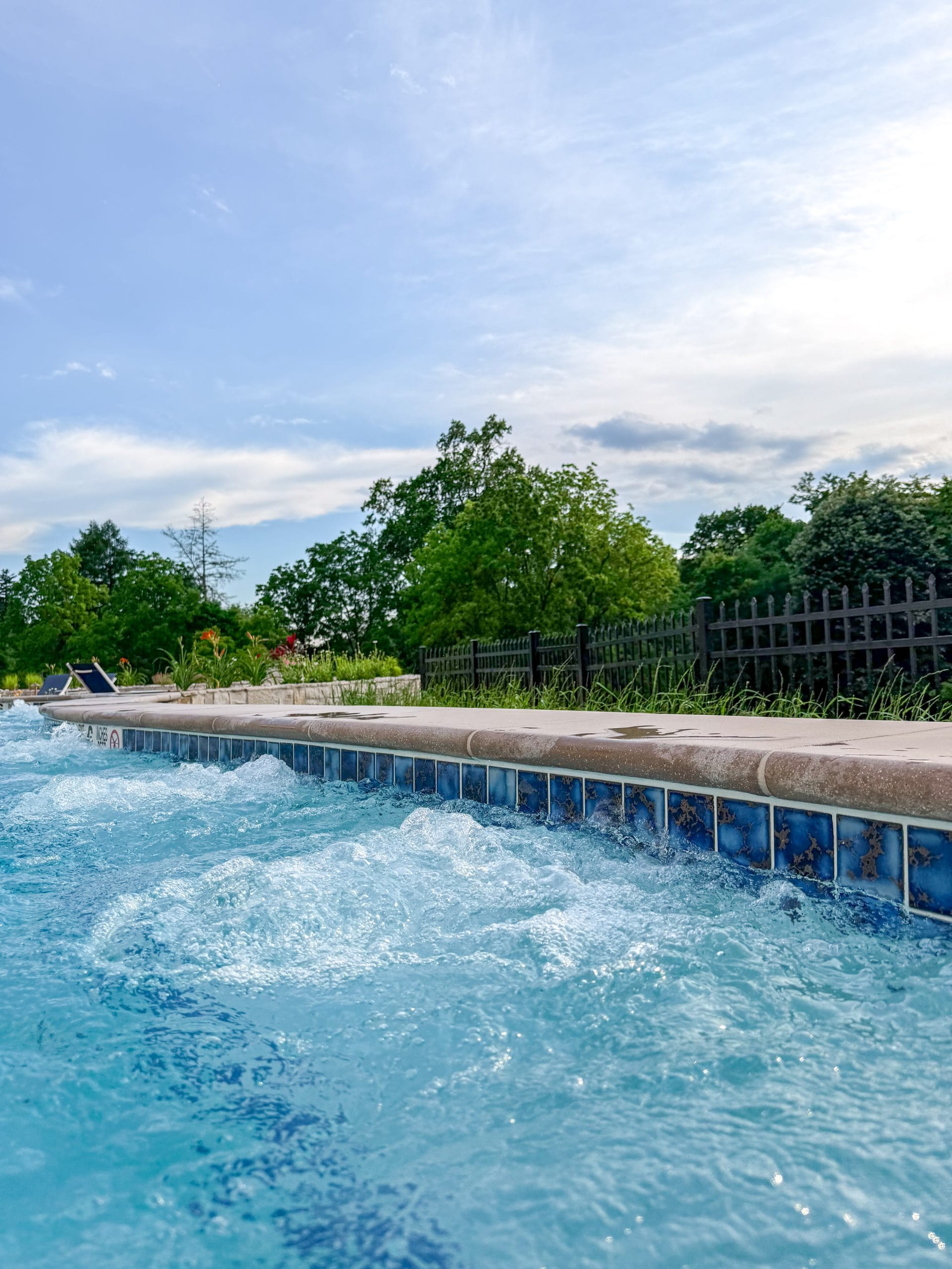 A close-up view of bubbling water in a swimming pool with blue tile trim at Miss NY Mirbeau Inn, surrounded by greenery, a black fence, and trees under a partly cloudy sky.