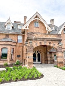 A large, historic brick building with arched stone entryway, decorative trim, multiple gabled roofs, and lantern-style lights—Miss NY Mirbeau Inn stands surrounded by neatly landscaped greenery and a paved walkway.