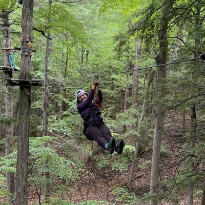 A person wearing a helmet and harness happily glides on a zipline through a lush, green forest, surrounded by tall trees and leafy branches. The forest floor is covered in fallen leaves and a wooden platform is visible below.