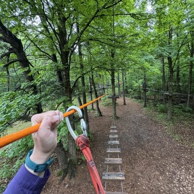 A person holds a safety clip attached to an orange cable while crossing a wooden rope bridge in a forest adventure park, surrounded by green trees and a dirt path below.