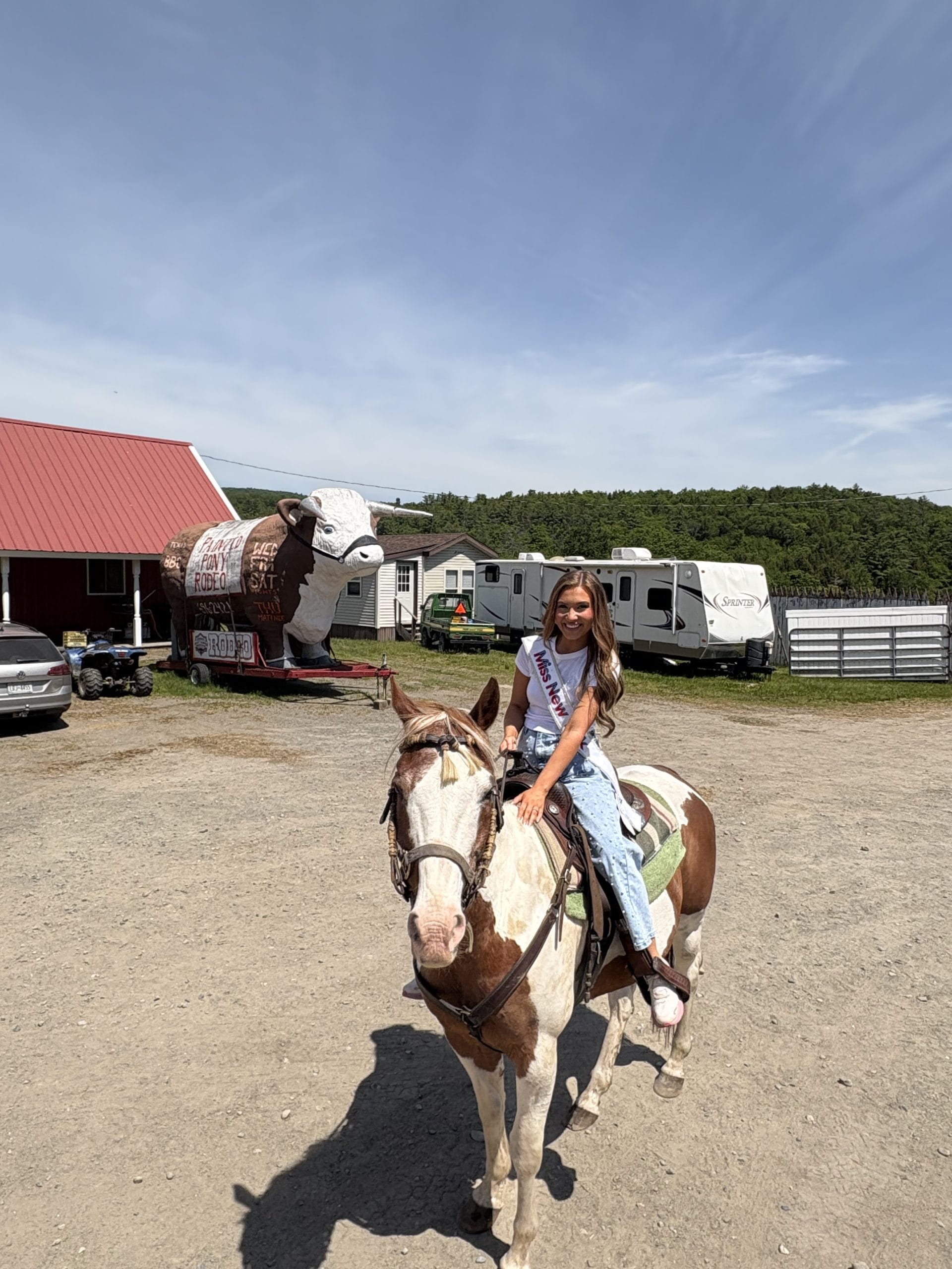 A young girl wearing a sash and crown sits on a brown and white horse at a fairground in the Lake George Area by Rail, with RVs, a red barn, and a large cow statue in the background under a blue sky.