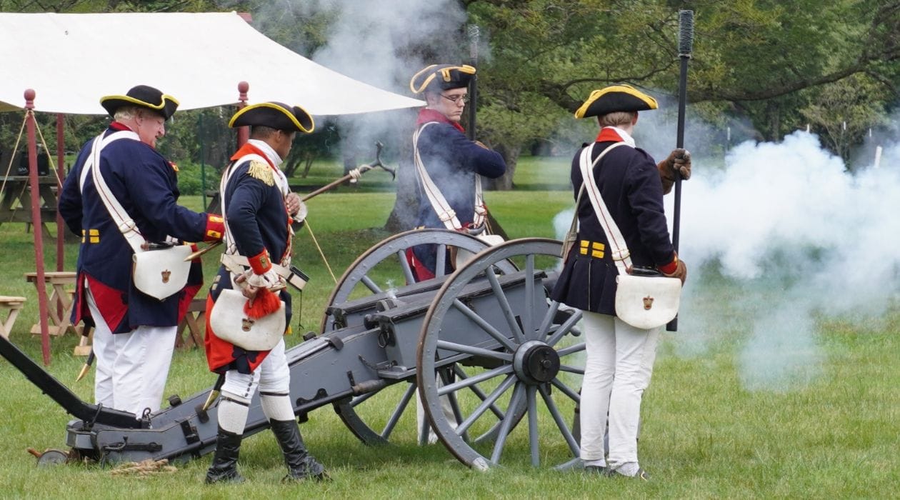 Four men in historical military uniforms fire a cannon outdoors at RevCon 2026, with smoke rising from the barrel. They stand on grass near a tent, reenacting a historical event.