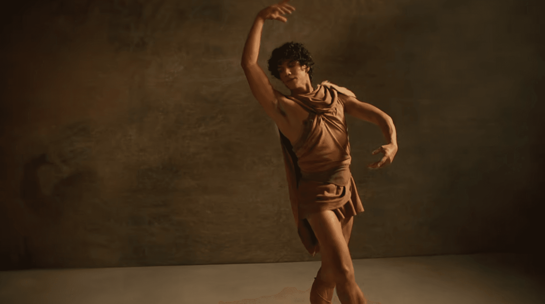 A male ballet dancer in a brown, ancient Greek-style costume performs gracefully against a dark studio backdrop, capturing the elegance and movement celebrated during the American Ballet Theatre Summer Season.