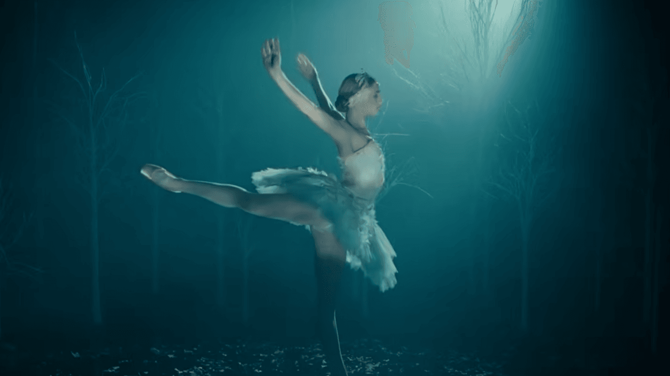 A ballerina in a white tutu performs an arabesque under a spotlight in a dark, misty forest with bare trees, evoking the enchanting artistry of the American Ballet Theatre Summer Season.