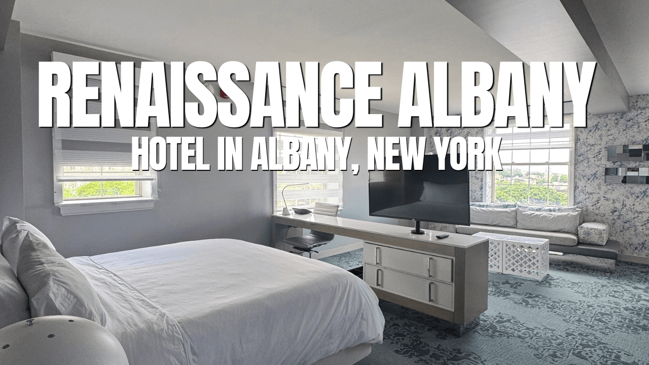 A modern hotel room with a large bed, TV, and seating area by the windows. White and pastel decor. Text reads: Renaissance Albany Hotel in Albany, New York—perfect for relaxing after a day of exploring the city or enjoying Hudson River Cruises.