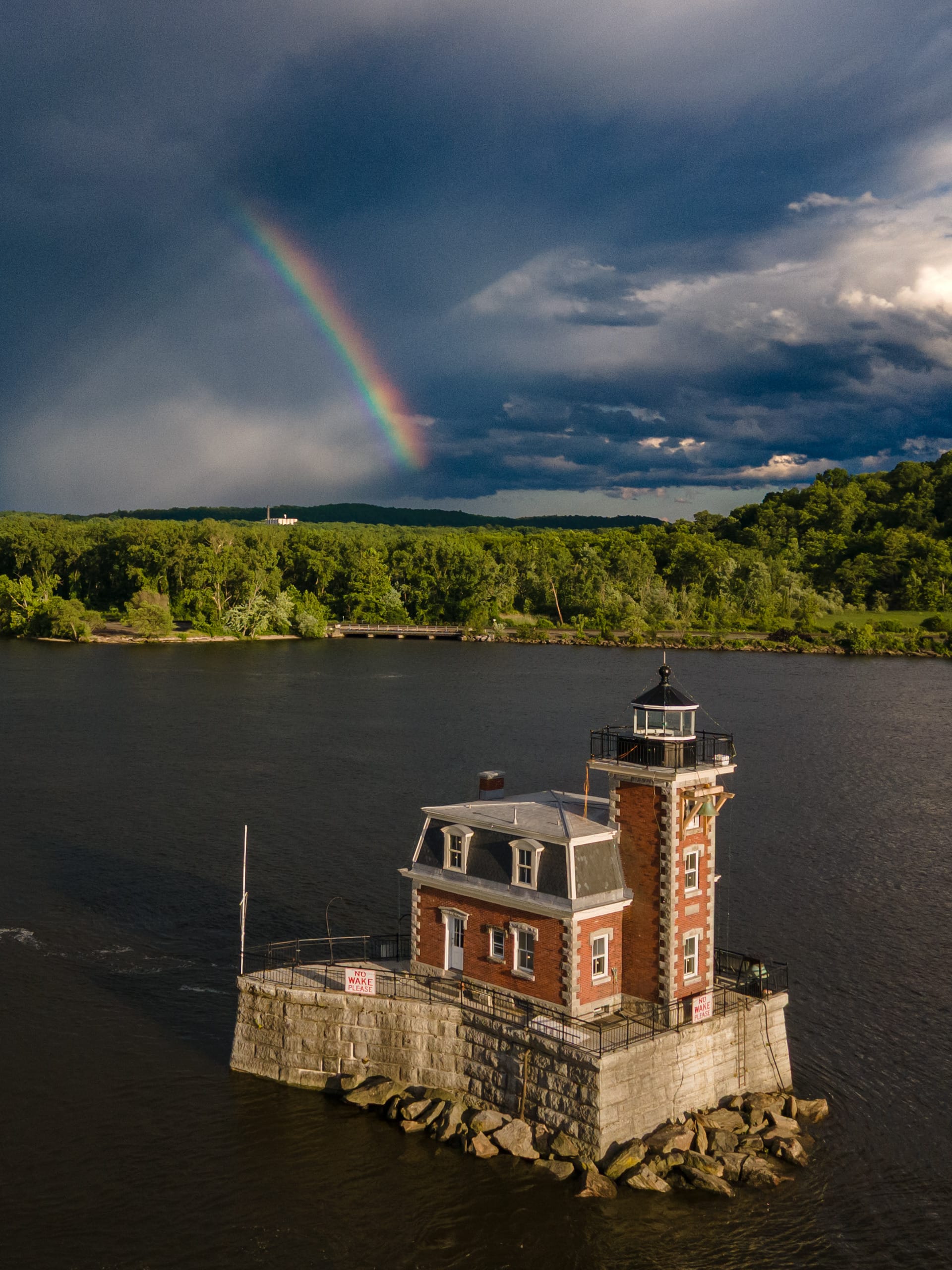 A small brick lighthouse sits on a river with green trees in the background. A vivid rainbow, reminiscent of the vibrant energy of Osheaga, arcs across a dramatic, cloudy sky.