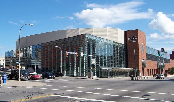 A modern glass and brick arena sits at a busy city intersection with cars and traffic lights—a must-see stop on any Rochester overnight itinerary. The building is labeled Blue Cross Arena near the top, set against a blue sky with scattered clouds.